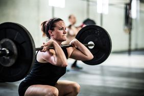 White woman wearing a black shorts and tank is performing a squat lifting a barbell in a gym setting. Her face is concentrated, looking forward with her lips pressed together