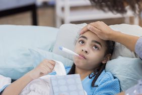 young child with thermometer in her mouth experiencing fever while laying down on a couch and holding a box of tissues