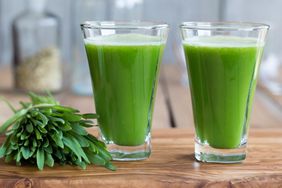 Two wheatgrass shots (green liquid in clear shot glasses) on a wood cutting board. A bundle of wheatgrass sits besides the shots