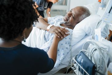 older adult lying in hospital bed while loved one holds their hand