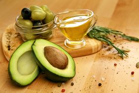 A bowl of olives and a halved avocado sit on a wood cutting board, along with a glass cup of oil. 