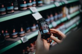 Over the shoulder view of woman browsing through medical products and reading the label on a bottle of supplements in front of the shelves in a pharmacy.