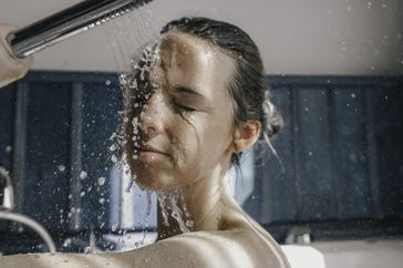 Young woman taking a warm shower in a sunlit bathroom in the morning, pouring water on her face. 