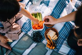 Young family eating carrots, celery, blueberries, crackers and cheese sticks while relaxing on picnic blanket in the park on a sunny day