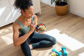 Shot of athletic woman eating a healthy fruit bowl while sitting on floor in the kitchen at home