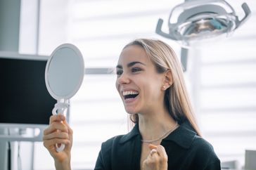 A blonde woman looking at her teeth in a handheld mirror at the dentist's office