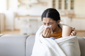 Sick young woman covered in blanket blowing her running nose in a tissue while sitting on couch in living room