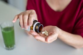 Woman pouring out supplement capsules into her hand; green juice in the background