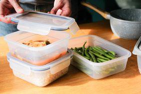 Person putting a lid on a plastic container of leftover food; three containers on a wooden counter top with a pot in the background
