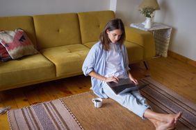 young woman typing at her laptop sitting on the floor at home with a cup 