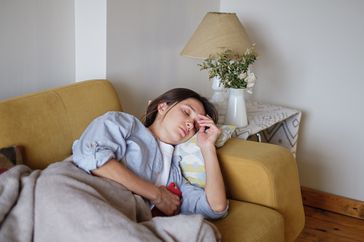 Young brunette woman feel sick and rest at home during the day under the plaid on the sofa scrolling her smartphone