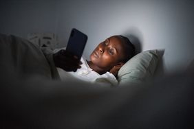 Young African American female addicted to devices lying in bed with hand under head and browsing mobile phone at night
