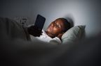 Young African American female addicted to devices lying in bed with hand under head and browsing mobile phone at night