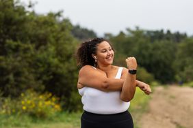 A smiling young woman stretches her arm during her exercise outdoors.