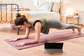 One brunette sportswoman standing in plank position on yoga mat training online via tablet in her bedroom