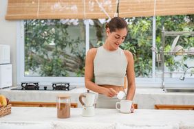 Smiling woman is adding a protein powder in a cup of coffee in the kitchen