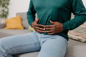 A man sitting on a sofa in his living room, holding his stomach due to digestive problems
