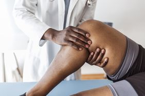 Closeup side view of early 30's unrecognizable doctor examining a knee of a woman during an appointment. The doctor is gently touching the tendons around the knee and the knee cap and trying to determine the cause of pain.