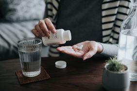 close up of a woman holding a pill bottle, pouring pills into palm of hand with a glass of water on the side