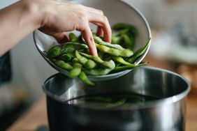 A person standing at the gas stove and putting edamame beans into boiling water.