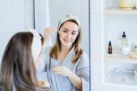 Young woman in bathrobe looking in the mirror, holding dropper with hyaluronic acid serum close to face and smiling