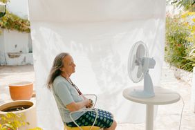 An gray haired woman sits outside in front of an electric fan on a hot day.
