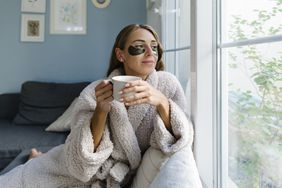 Woman wearing bathrobe, wearing under-eye patches, holding coffee cup, looking through window
